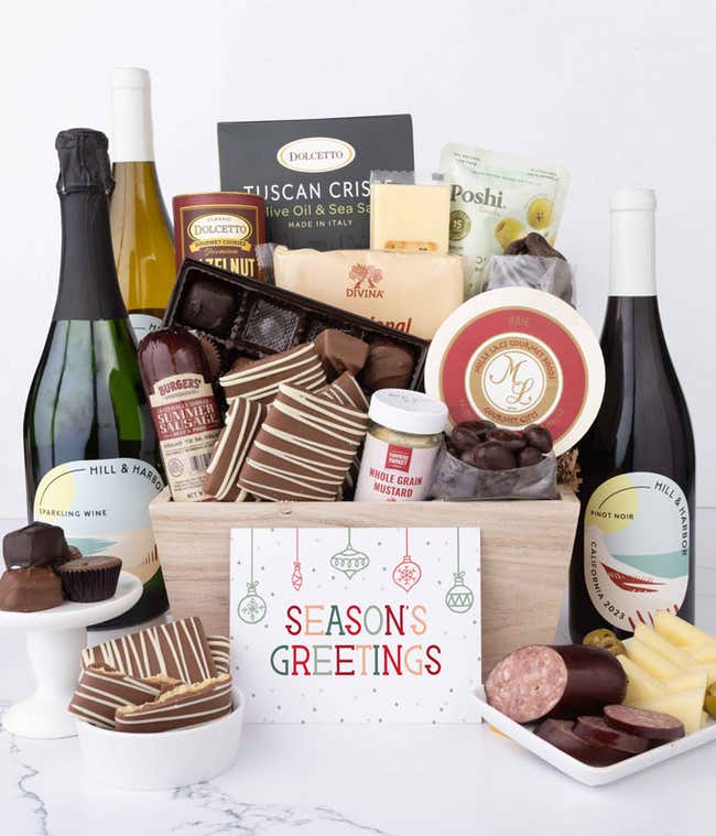 A wooden basket filled with chocolates, summer sausage, cheeses, crackers, crisps, mustard, and snacks, with displays of chocolate treats, sliced sausage, olives, and cheese arranged with 3 bottles of wine and Season's Greetings message.