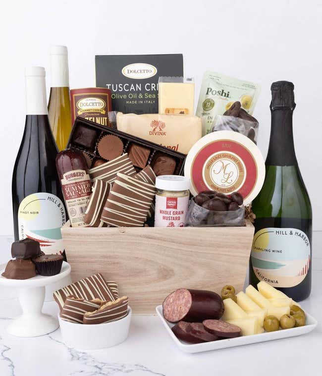 A wooden crate filled with gourmet snacks including sausage, assorted cheeses, crackers, olives, mustard, chocolate almonds, pretzels, graham crackers, assorted chocolates, and wafers, with 3 bottles of wine and an Easter card.