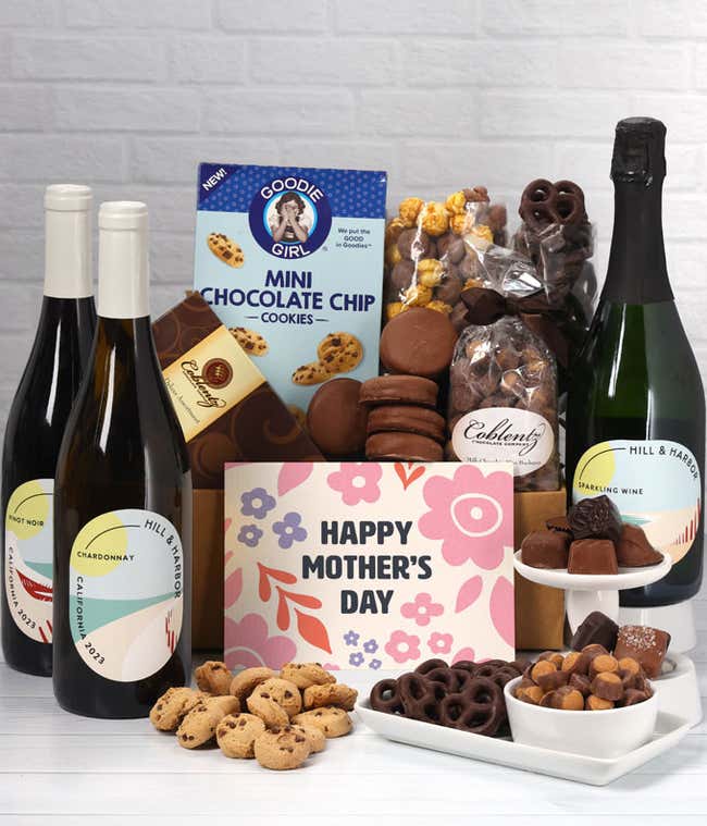 Three bottles of wine - red, white &amp; sparkling, sit beside chocolate-covered pretzels, mini chocolate chip cookies, caramel popcorn, assorted chocolates, mini buckeyes, chocolate covered Oreos, and a Happy Mother's Day card.