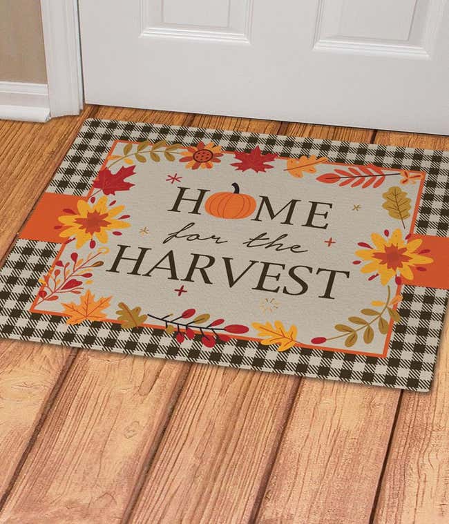 Home for the Harvest doormat with autumn-themed design, featuring fall leaves and a pumpkin, bordered by a black and white buffalo plaid pattern.