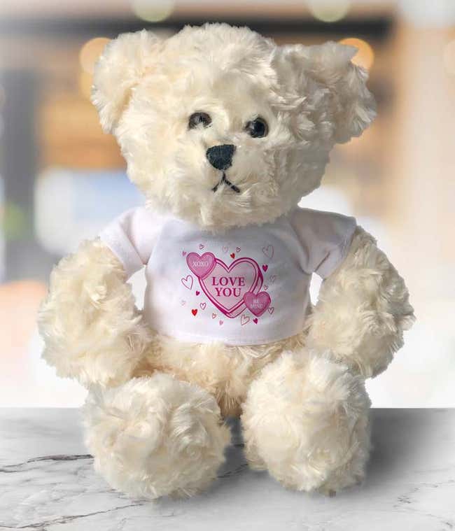 Fluffy cream-colored teddy bear wearing a white shirt featuring a pink heart design with the words 