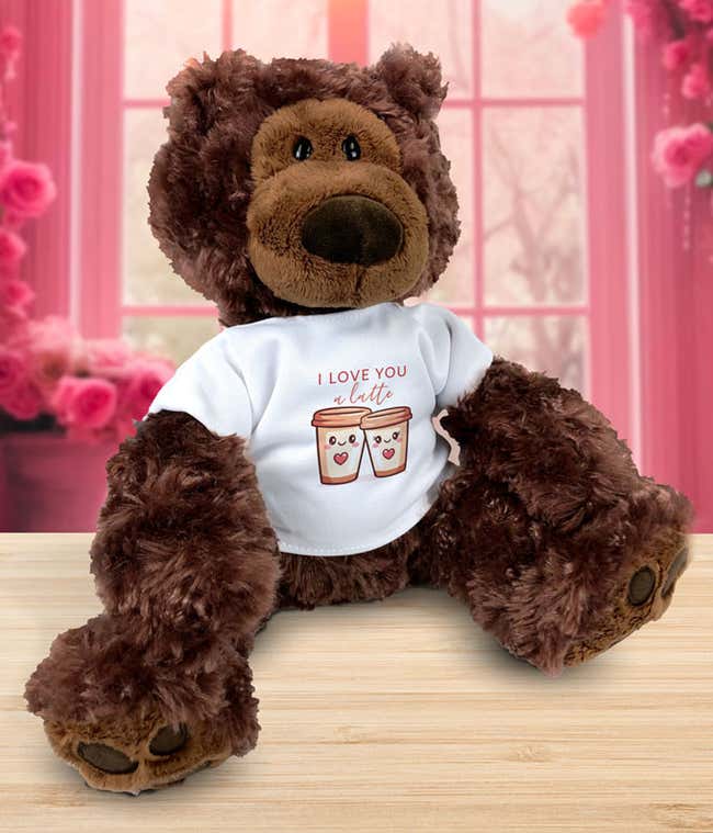 Plush brown teddy bear sitting on a wooden surface, wearing a white T-shirt that reads &lsquo;I love you a latte&rsquo; with an illustration of two smiling coffee cups, set against a pink, rose-filled background.
