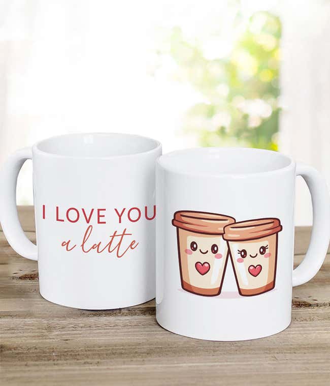 wo white coffee mugs, one reading &lsquo;I love you a latte&rsquo; and the other featuring an illustration of two smiling coffee cups with heart details, displayed on a rustic wooden surface.
